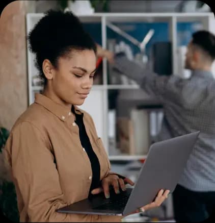 A woman standing and using a laptop, focused on her screen, while a man organizes files on a shelf in the background.